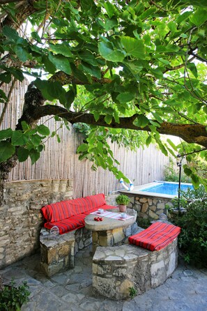 A stone patio with a red-striped sofa and cushions, a small table with a potted plant, and a view of a swimming pool in the background. - Villa Lithos (Faistos)