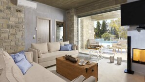 A modern living room with a stone wall, a beige sofa, a wooden coffee table, and a large window overlooking a pool. - Kouros Villa (Faistos)