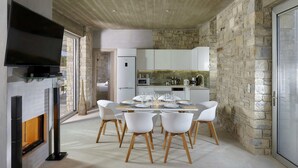 A modern dining area with a large table set for six, surrounded by white chairs with wooden legs. A flat-screen TV is mounted on the wall above the table. - Kouros Villa (Faistos)