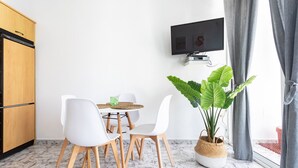 Een moderne kamer met een eettafel, witte stoelen, een aan de muur bevestigde televisie, een grote potplant en een houten kast.