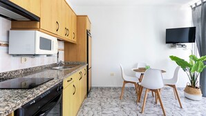 Een kleine keuken met een magnetron, spoelbak en houten keukenkastjes. Er is een eethoek met witte stoelen en een tafel, en een potplant.