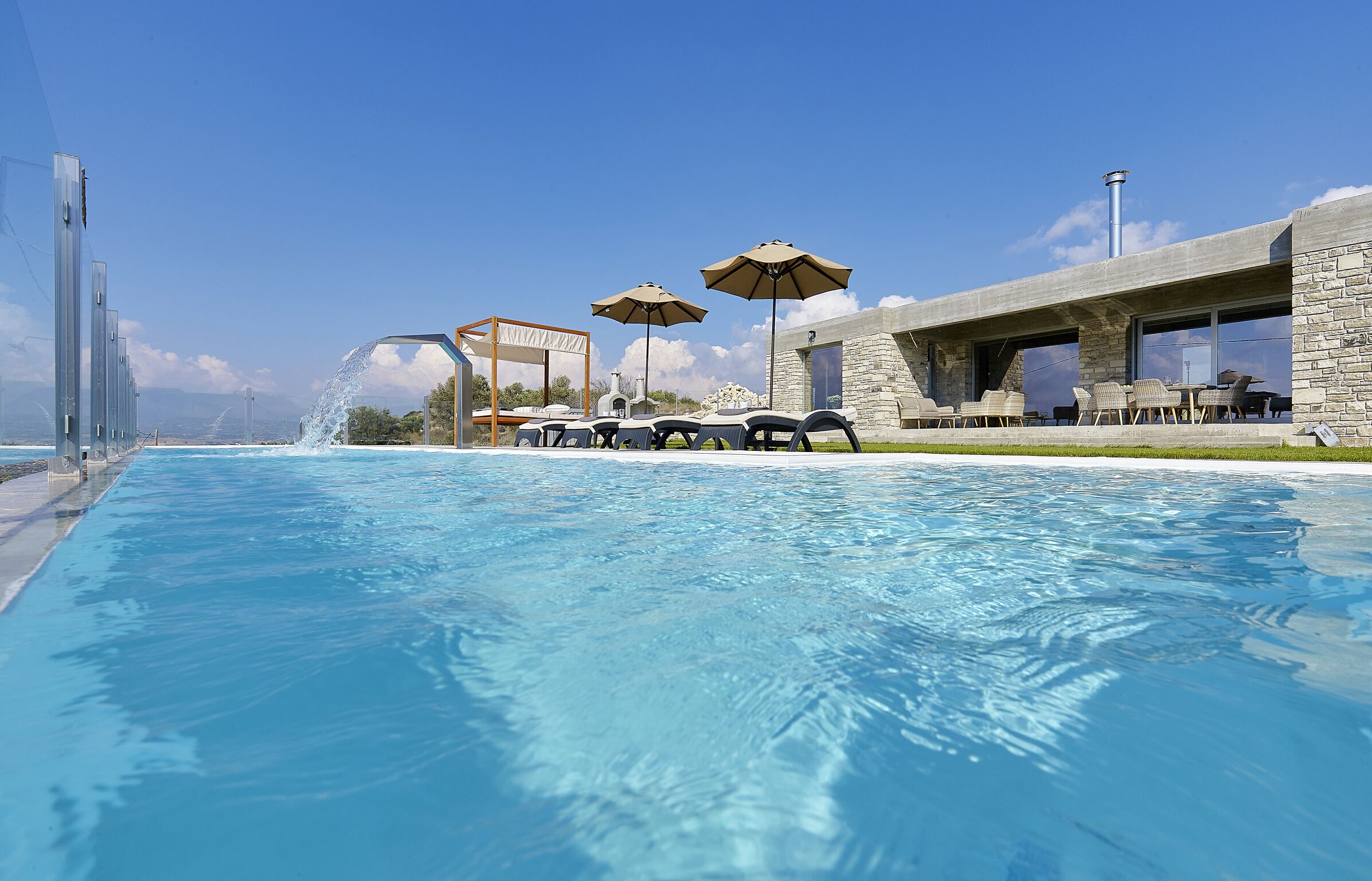 A swimming pool with lounge chairs and umbrellas, a stone building with large windows, and a clear blue sky.
