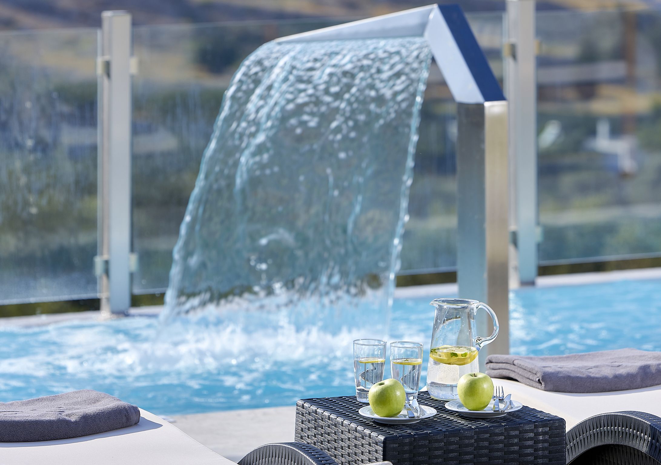 A poolside setting with a waterfall feature, a table with glasses and a pitcher, and a towel.