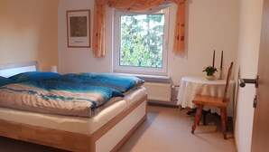 A bedroom with a bed, a window with curtains, a radiator, a small table with a plant, and a chair.