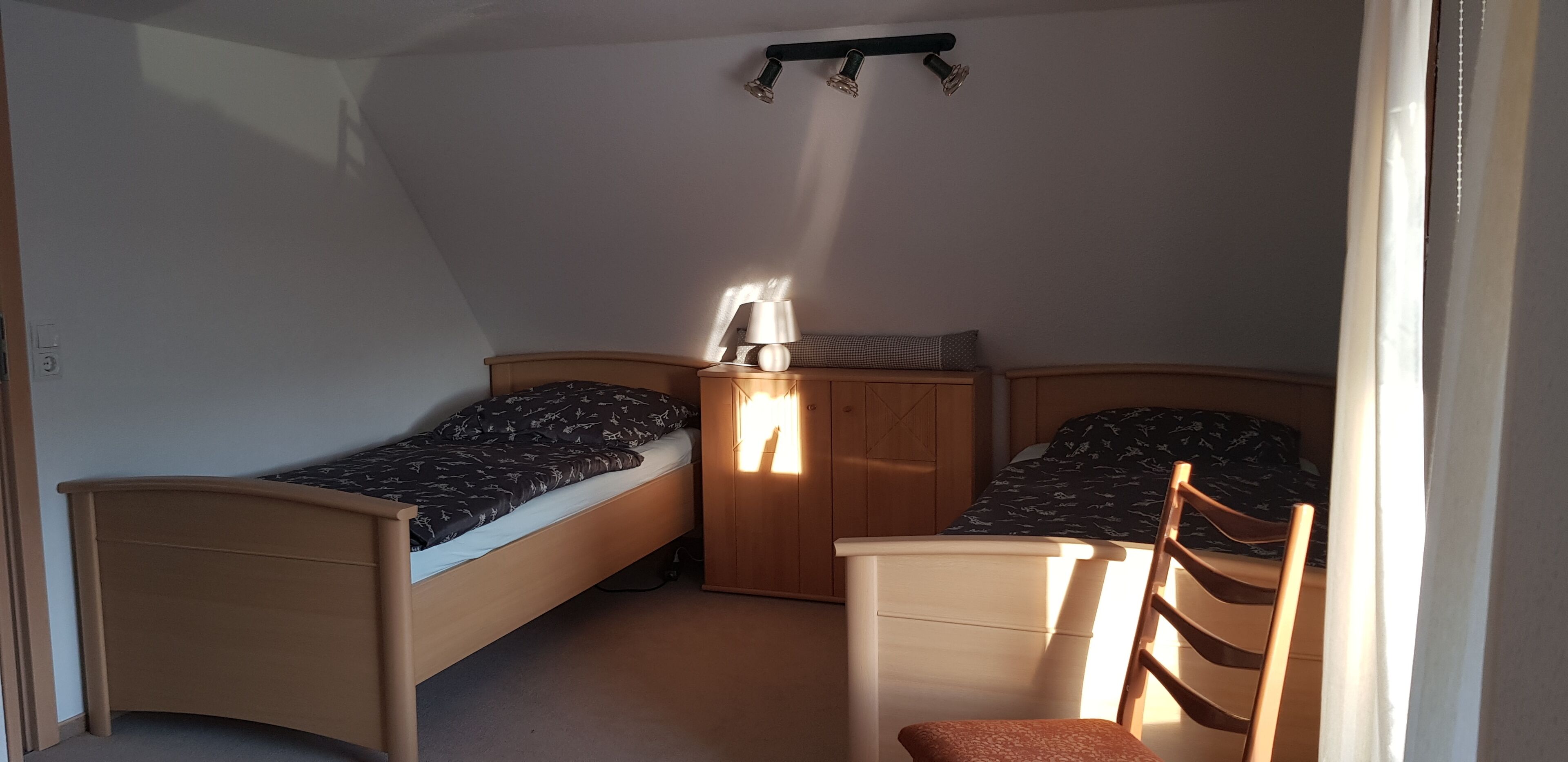 Un dormitorio pequeño y luminoso con dos camas individuales, una silla de madera y una mesita de noche con lámpara.