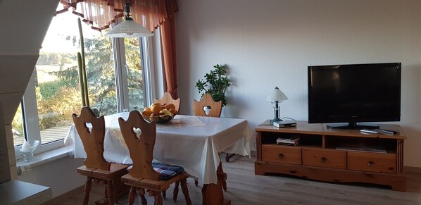 Ein Essbereich mit einem für zwei Personen gedeckten Tisch, einem Fernseher auf einem Holzständer und einem Fenster mit Vorhängen.