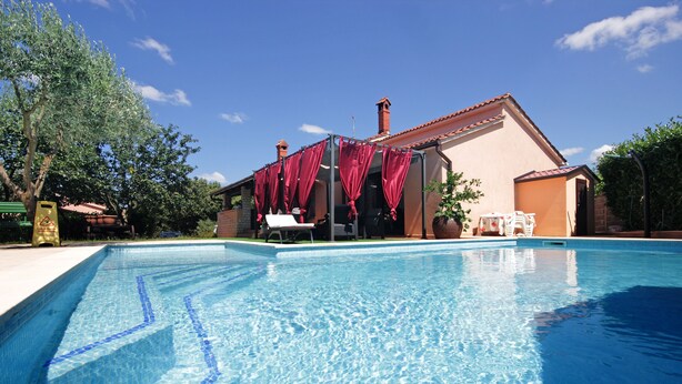 A swimming pool with a clear blue waterline, a covered outdoor seating area, and a building with a tiled roof in the background.