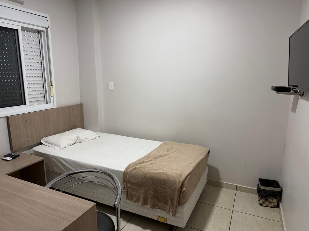 A small, single-bed room with a desk, chair, and a window with blinds.