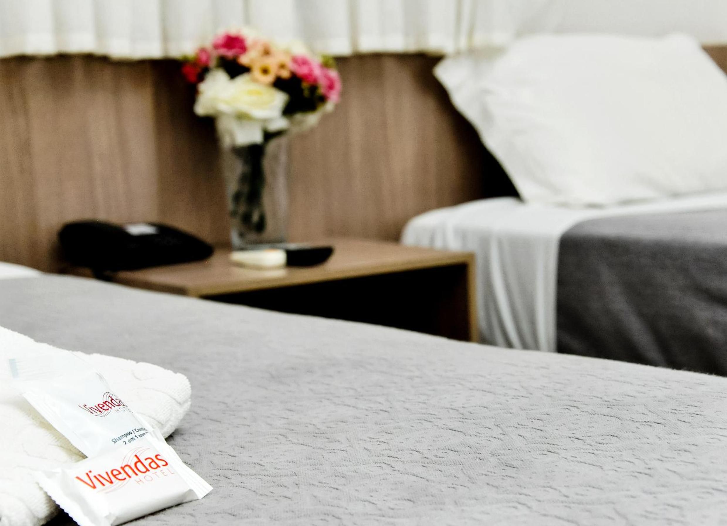 Una habitación de hotel con una cama bien hecha, una mesita de noche con un jarrón de flores y una etiqueta identificativa del hotel a la vista.