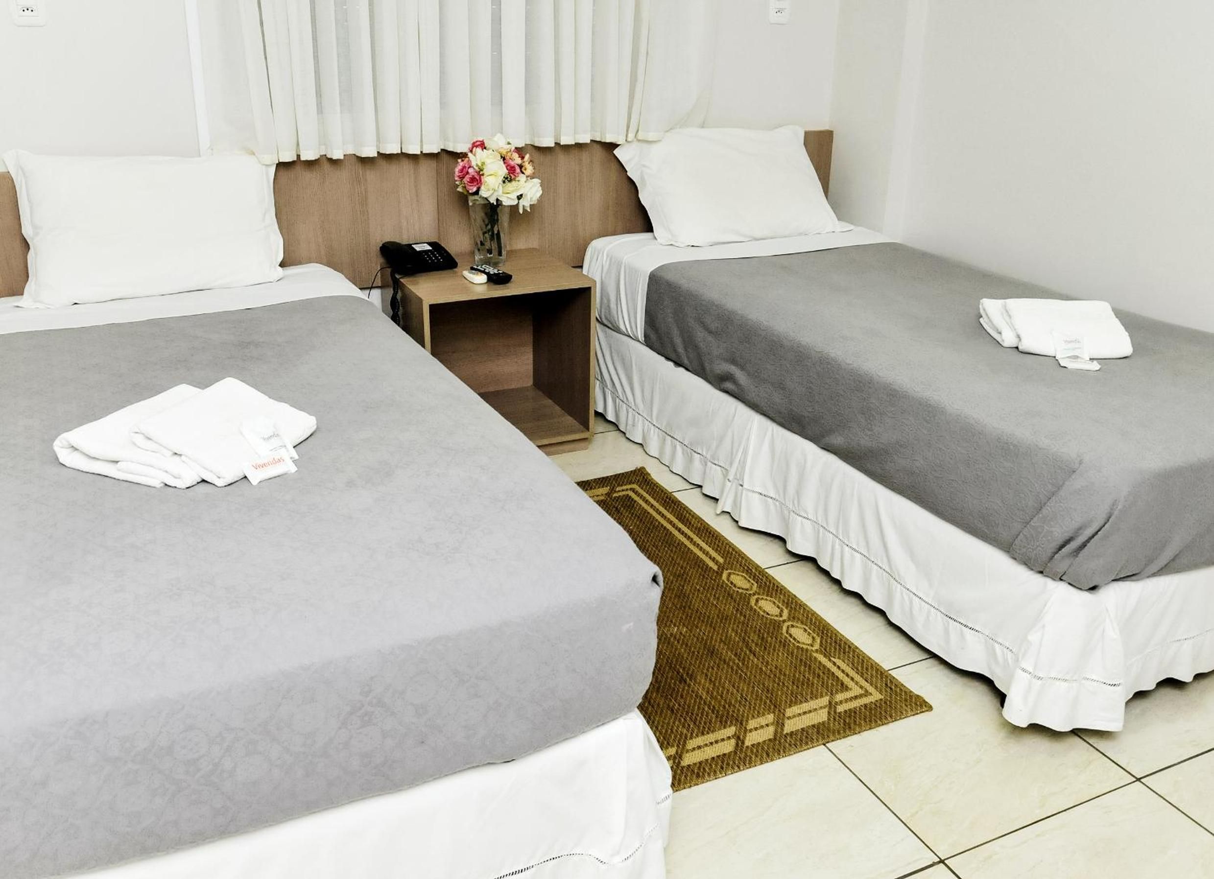A hotel room with two beds, a nightstand, a vase with flowers, and a rug.