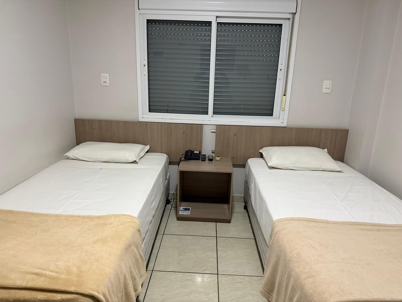 Una habitación con dos camas, una mesita de noche pequeña con un teléfono y una ventana con persianas.