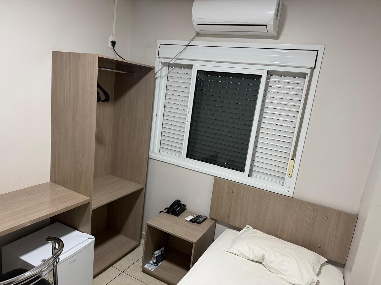A compact hotel room with a bed, a bedside table with a phone and remote, a small desk with a chair, and a window with blinds.