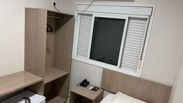A compact hotel room with a bed, a bedside table with a phone and remote, a small desk with a chair, and a window with blinds.