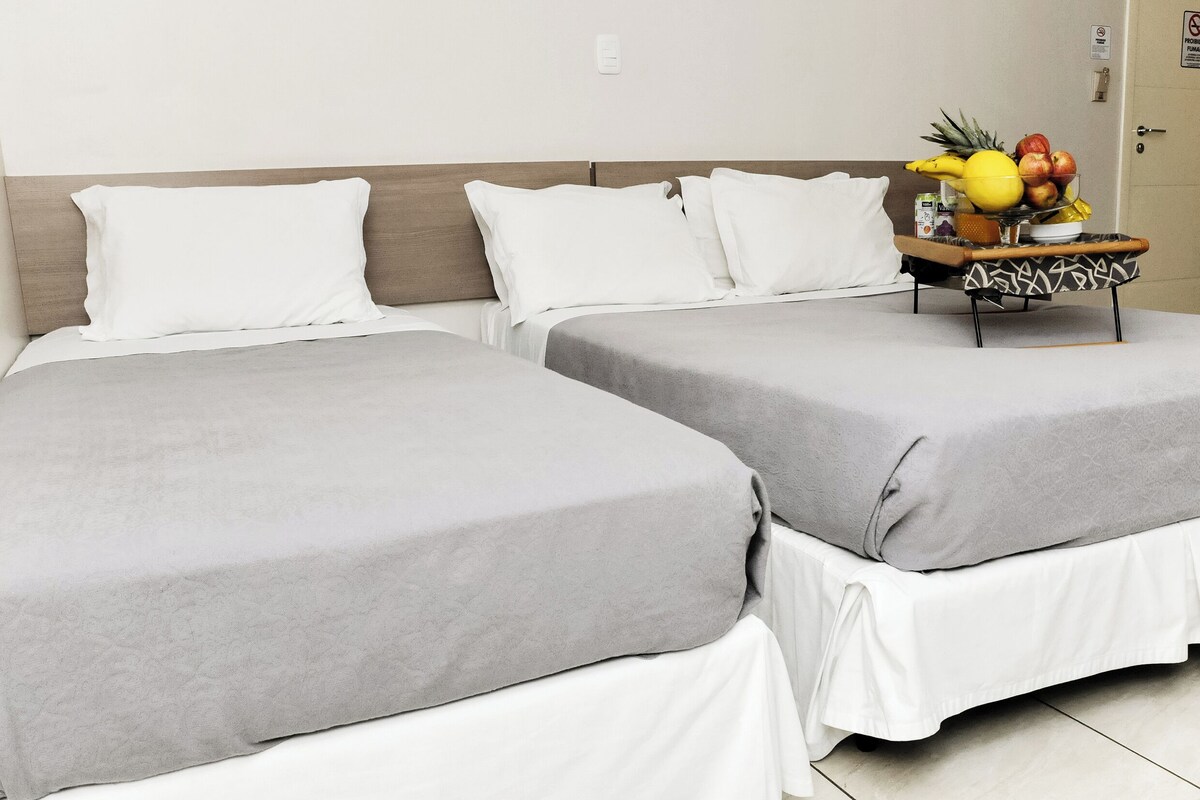 Habitació d'hotel amb dos llits, cadascun amb estovatge gris i naxes blanques. Hi ha una taula petita amb un plat de fruita i una ampolla.