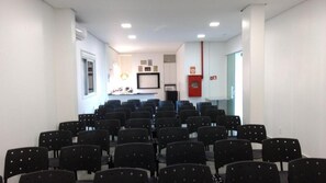 Una sala de conferències amb files de cadires negres, parets blanques i una zona de cuina al fons.