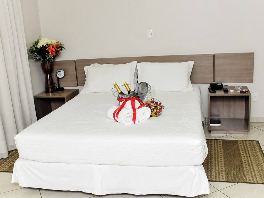 Een hotelkamer met een net opgemaakt bed, een nachtkastje met een lamp, een bijzettafeltje met een telefoon en een glas, en een decoratieve vaas met bloemen.