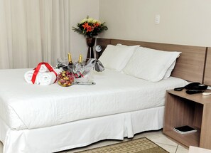 A neatly made bed with white linens, a bedside table with a phone, a small tray with a bottle, glasses, and a wrapped item. - Vivendas Hotel (Erechim)
