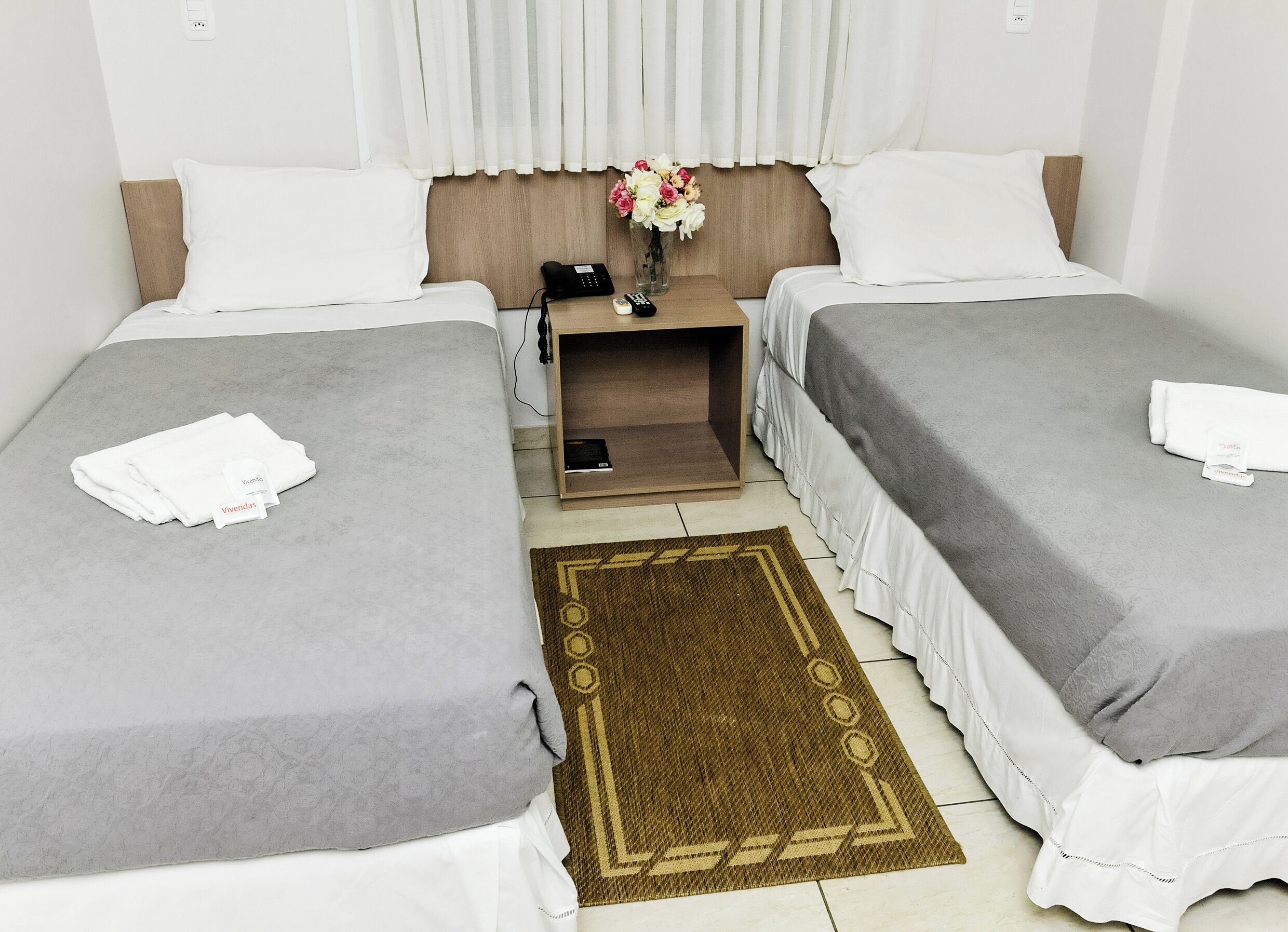 Habitación de hotel con dos camas, una mesita de noche con teléfono, un florero con flores y una alfombra marrón.