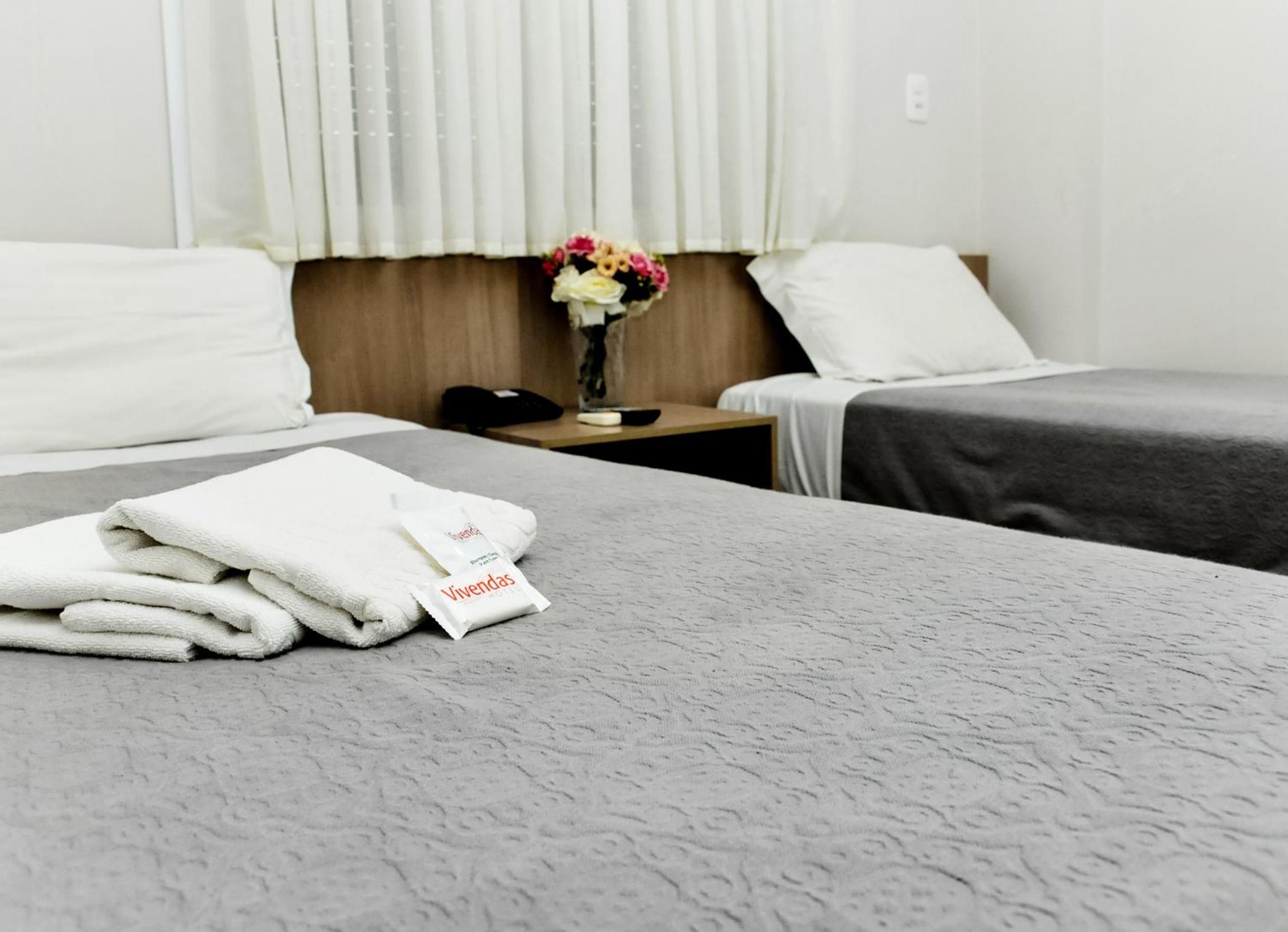 A hotel room with two beds, a nightstand with a phone, a vase with flowers, and a package labeled 'Vivendas'.