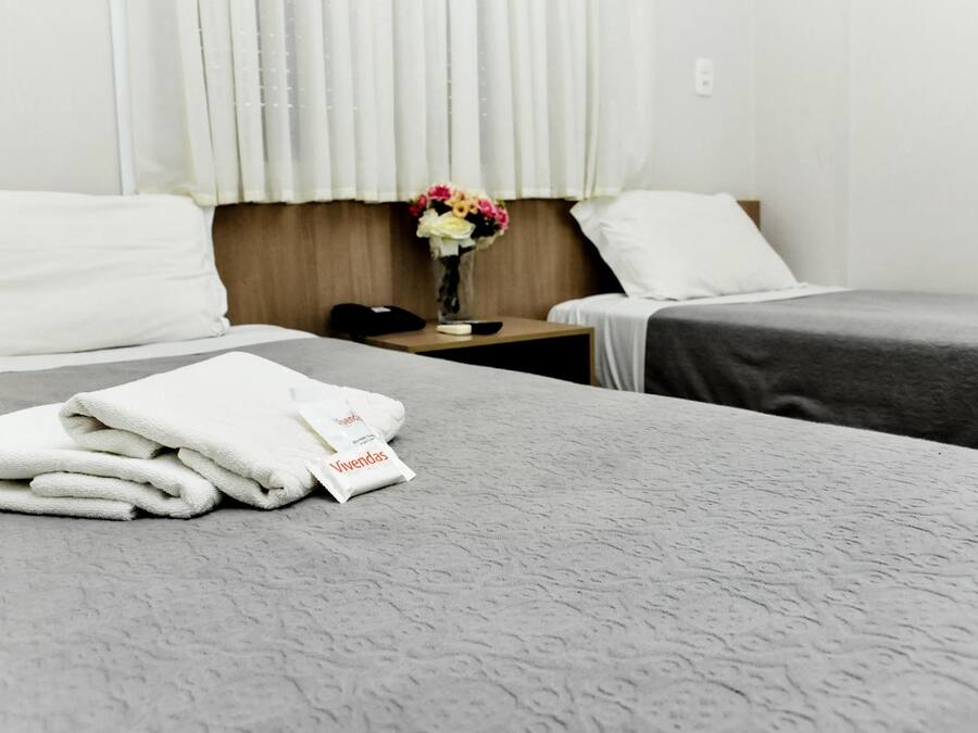 Habitación de hotel con dos camas, una mesita de noche con un teléfono, un jarrón con flores y un paquete etiquetado 'Vivendas'.