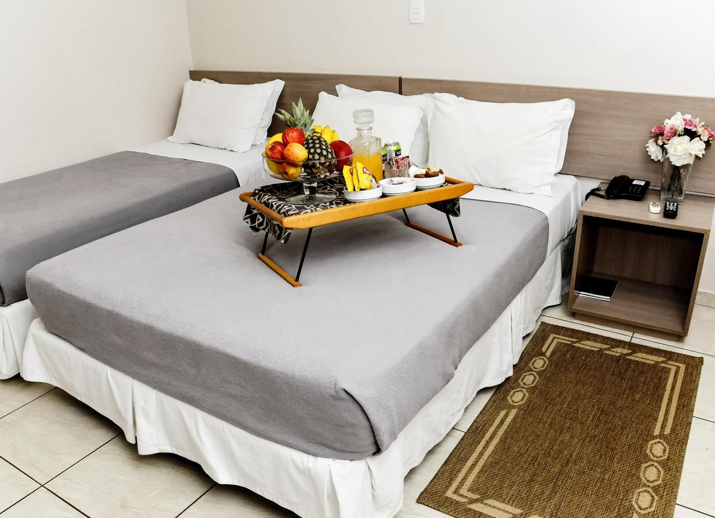 Quarto de hotel com duas camas, uma mesinha de cabeceira com telefone, um vaso com flores e uma bandeja com frutas e uma garrafa.