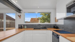 Una cocina moderna con encimera de madera, armarios blancos, horno empotrado y ventana con vista a una casa y árboles.