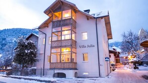 Un edificio a più piani, Villa Julia, con un balcone, circondato da alberi e montagne innevate.