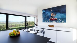 Un espace de repas moderne avec une grande table, des chaises et un saladier rempli de fruits. Un grand tableau orne le mur, et de larges fenêtres offrent une vue sur la campagne.