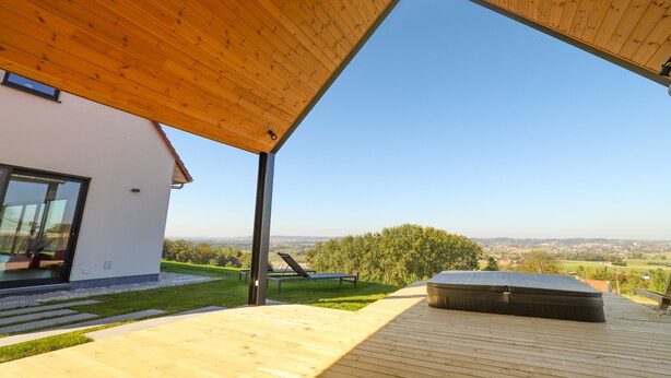 Une terrasse extérieure moderne, dotée d’un toit en bois, d’un jacuzzi et offrant une vue sur un paysage verdoyant et des bâtiments éloignés.