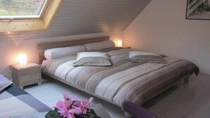 Een slaapkamer met een schuine kap, een groot bed met gestreepte lakens, nachtlampjes en een raam met uitzicht op bomen.