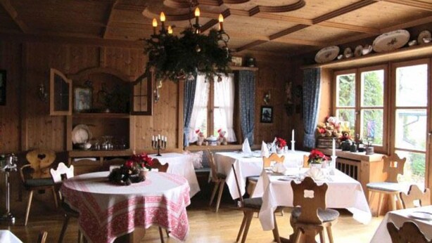 A rustic dining area with wooden tables and chairs, a chandelier, and a view of the outdoors through large windows. - Hotel & Restaurant Tonihof (Eschenlohe)