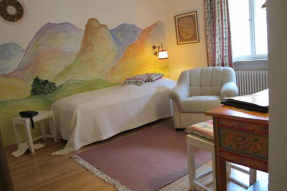 A bedroom with a bed, a chair, a desk, and a wall painting.