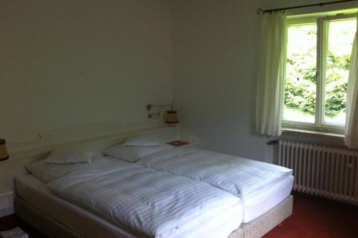 A bedroom with a double bed, a nightstand, a lamp, a window with curtains, and a radiator.