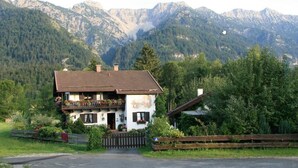 A two-story house with a balcony, surrounded by lush greenery and mountains in the background. - Hotel & Restaurant Tonihof (Eschenlohe)