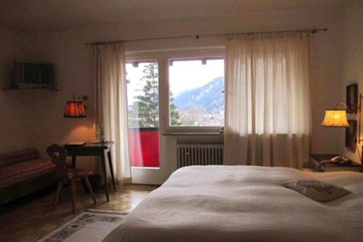 A hotel room with a bed, a desk, a chair, a window with curtains, and a view of mountains.