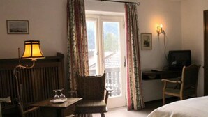 A hotel room with a bed, a desk with a computer, a chair, a lamp, and a window with curtains. - Hotel & Restaurant Tonihof (Eschenlohe)