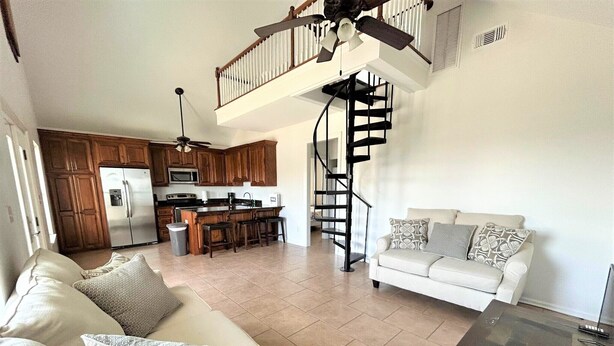 A spacious living area with a white sofa, patterned cushions, a glass coffee table, a kitchen with wooden cabinets, a built-in island, and a spiral staircase. - Blue Water Villa B - Coastal Realty Group (Grand Isle)