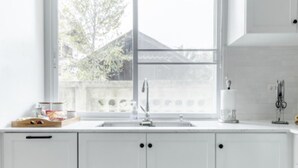 A kitchen with white cabinets, a sink, and a window with a view of a fence and trees. - Discover Chiang Mai from Cheerful Contemporary Cottage (Muang Chiang mai)