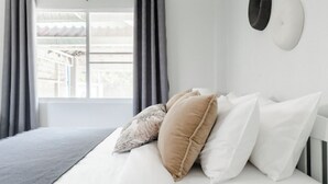 A neatly made bed with white and beige pillows, a gray throw blanket, and a window with gray curtains. - Discover Chiang Mai from Cheerful Contemporary Cottage (Muang Chiang mai)
