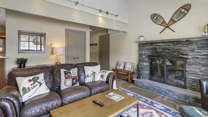 A living room with a leather sofa, a fireplace, and a stone wall. - Stowe Base Camp 21 at Stonybrook: 3 br, fireplace, a/c, decks, summer pool & community amenities! (Stowe)
