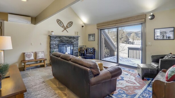 A spacious living room with a stone fireplace, a large sofa, and a view of a snowy landscape through a sliding glass door. - Stowe Base Camp 21 at Stonybrook: 3 br, fireplace, a/c, decks, summer pool & community amenities! (Stowe)