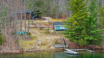 Una cabaña junto al lago con un muelle, rodeada de árboles y un pequeño puente.