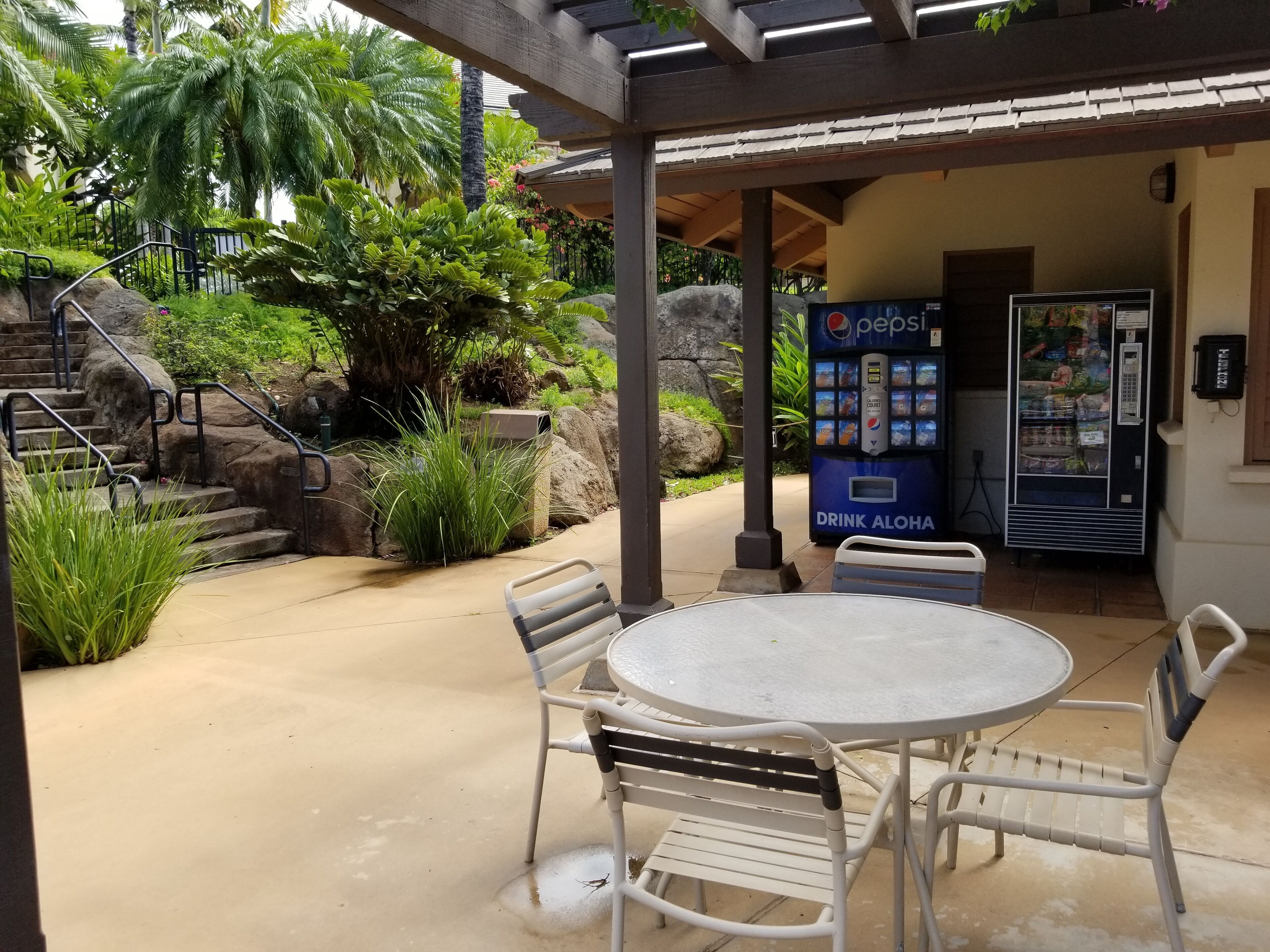 An outdoor patio with a round table and chairs, a Pepsi drink vending machine, and a stone staircase leading to a covered area.