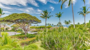 A tropical landscape with palm trees, a large tree with a broad canopy, and a view of the ocean. - Top Floor Spacious Ocean View Condo! Minutes to Beaches, A/C, Wifi, Pool (Kihei)