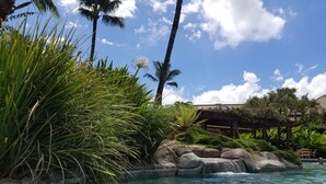 A clear pool surrounded by tropical vegetation and a wooden structure. - Top Floor Spacious Ocean View Condo! Minutes to Beaches, A/C, Wifi, Pool (Kihei)