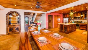 Un comedor amplio con una larga mesa de madera dispuesta para una comida, una cocina con electrodomésticos modernos y una escalera que conduce a otro nivel.