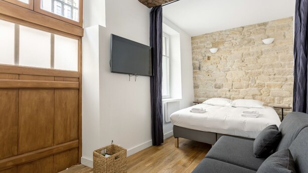 A bedroom with a stone wall, a bed, a grey sofa, a TV, and a wooden door. - Saint Paul, Marais History France Louis XIV &XV (Paris)