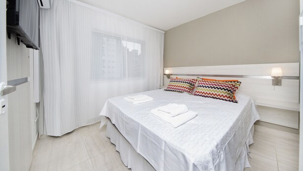 Quarto de hotel com uma cama grande, cortinas brancas, cabeceira com iluminação embutida e uma TV de tela plana na parede.
