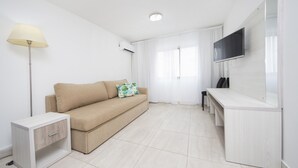 Uma sala de estar moderna e minimalista com um sofá bege, uma mesinha lateral, um abajur, uma TV de tela plana na parede e uma janela grande com cortinas translúcidas.
