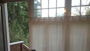 A room with a table and two chairs, a window with curtains, a wooden deck, and a view of trees.